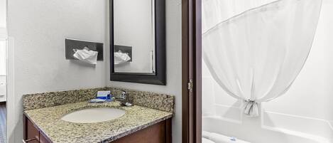Une salle de bain avec un lavabo sur un plan de travail en granit, un miroir et une baignoire blanche avec un rideau.