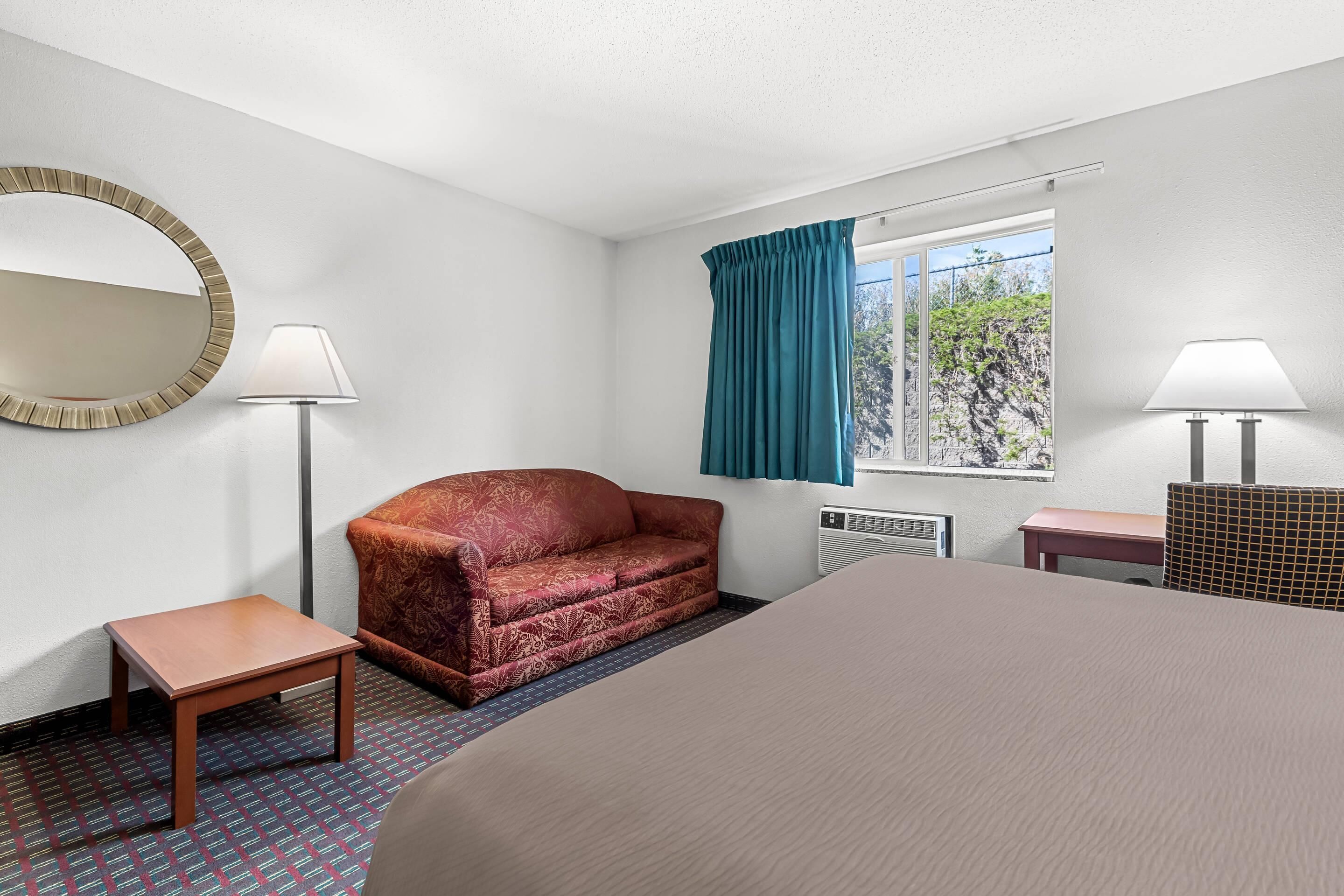 Una habitación de hotel con una cama, un sofá rojo estampado, una mesita, un espejo, dos lámparas y una ventana con cortinas.
