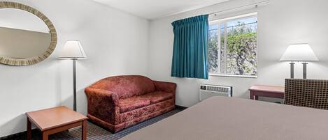 Una habitación de hotel con una cama, un sofá rojo estampado, una mesita, un espejo, dos lámparas y una ventana con cortinas.