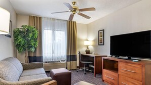 A hotel room with a sofa, ottoman, desk, television, and ceiling fan. - Comfort Inn & Suites Lees Summit - Kansas City (Lee's Summit)