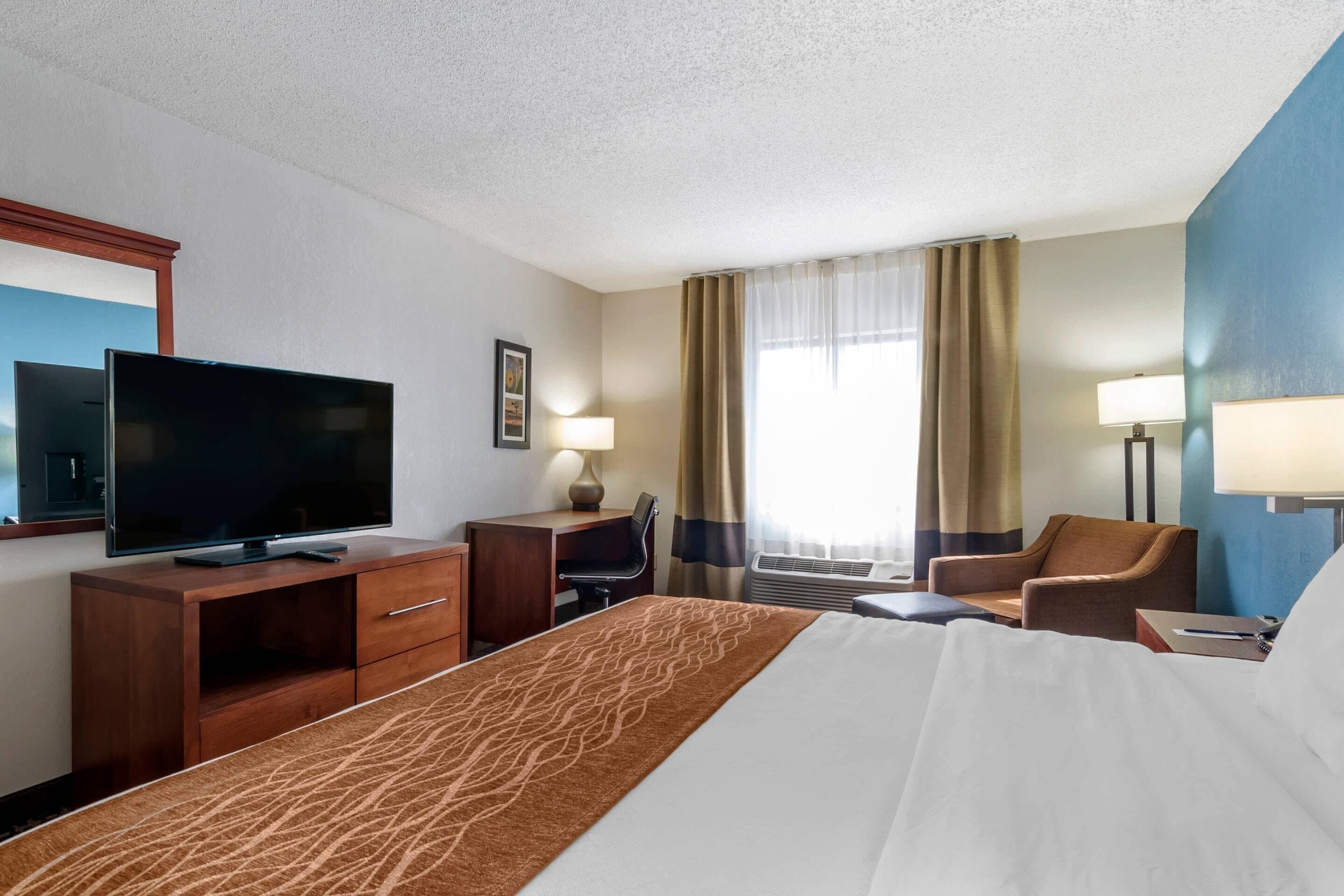 Ett hotellrum med en säng, ett skrivbord med en tv, en stol och ett fönster med gardiner.