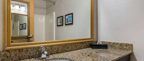 Un baño con encimera de granito, lavabo, espejo con marco dorado y dos cuadros en la pared.