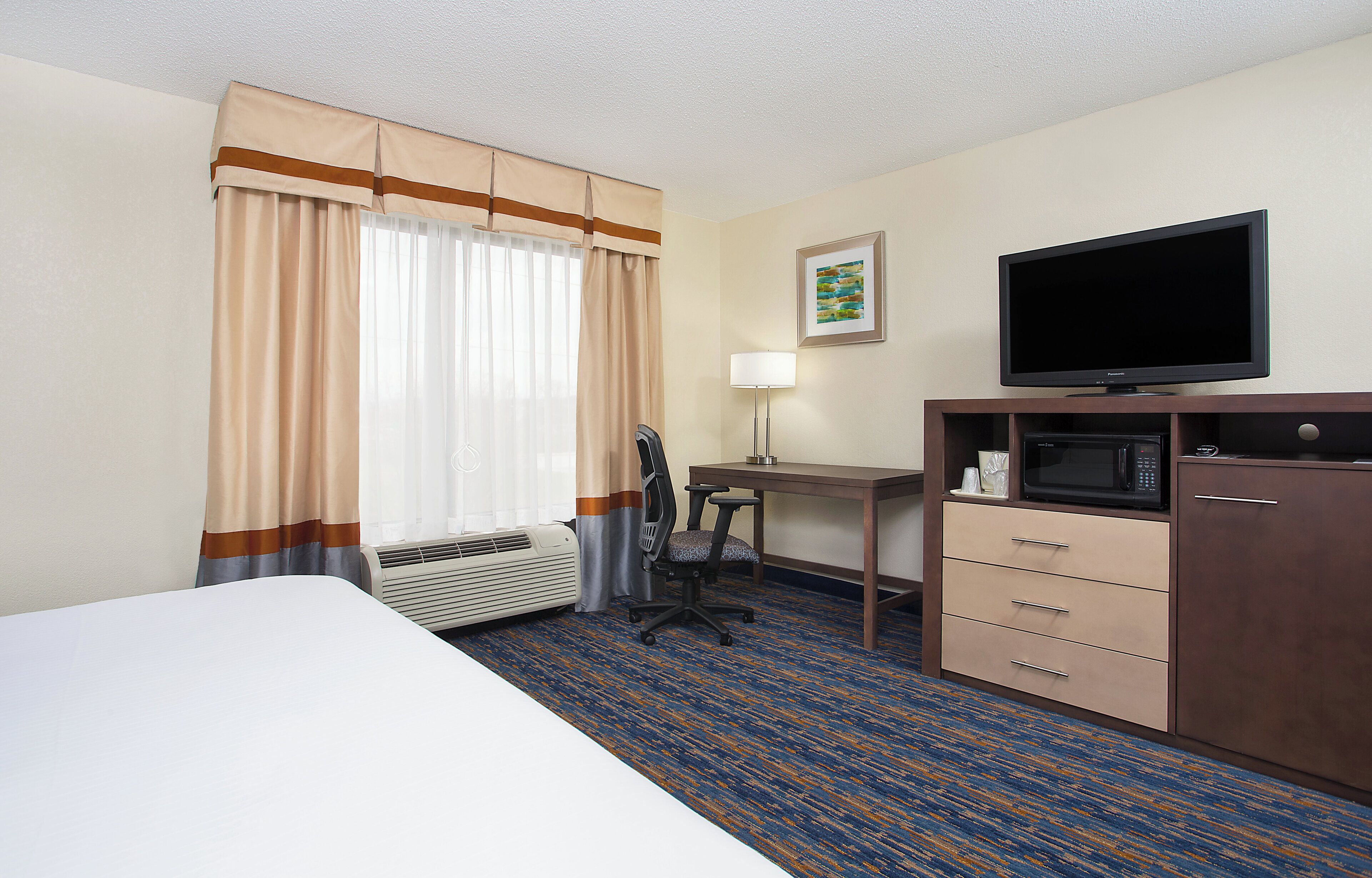 A hotel room with a bed, a desk with a computer, a television, a microwave, and a chair.
