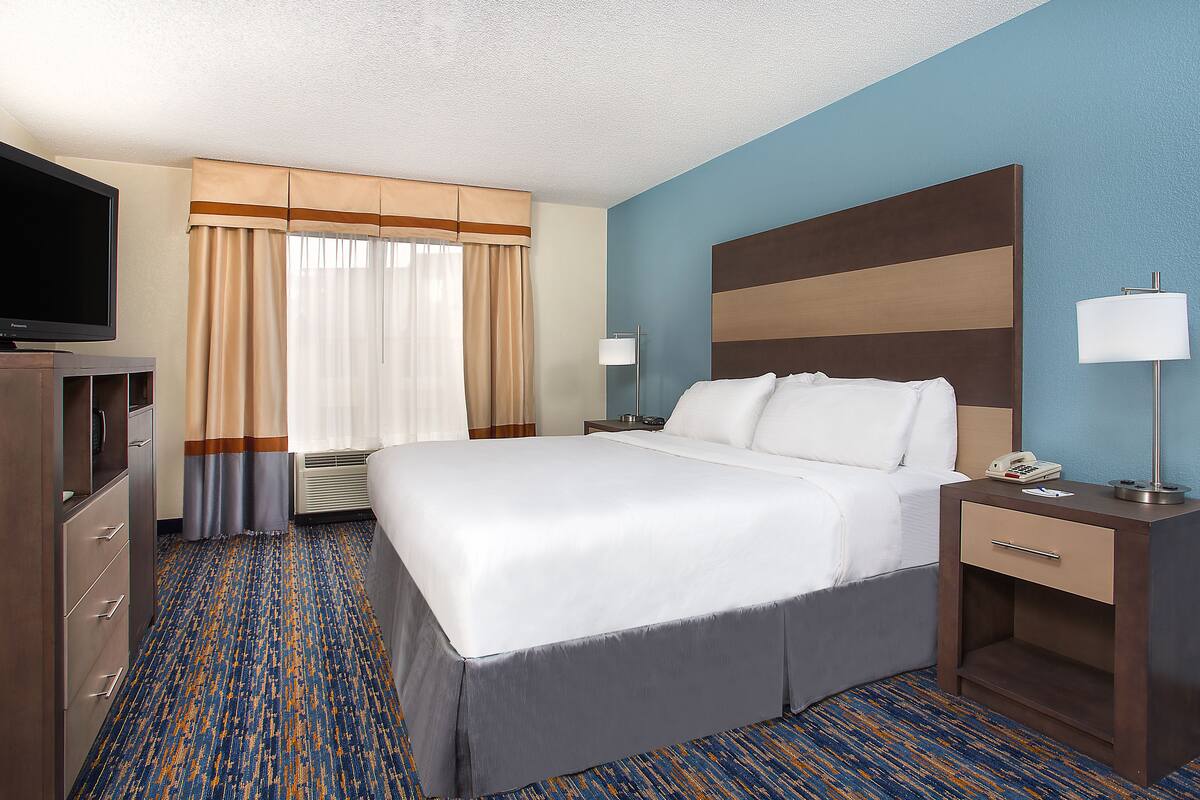 A hotel room with a large bed, a TV, a desk with a lamp, and a window with curtains.