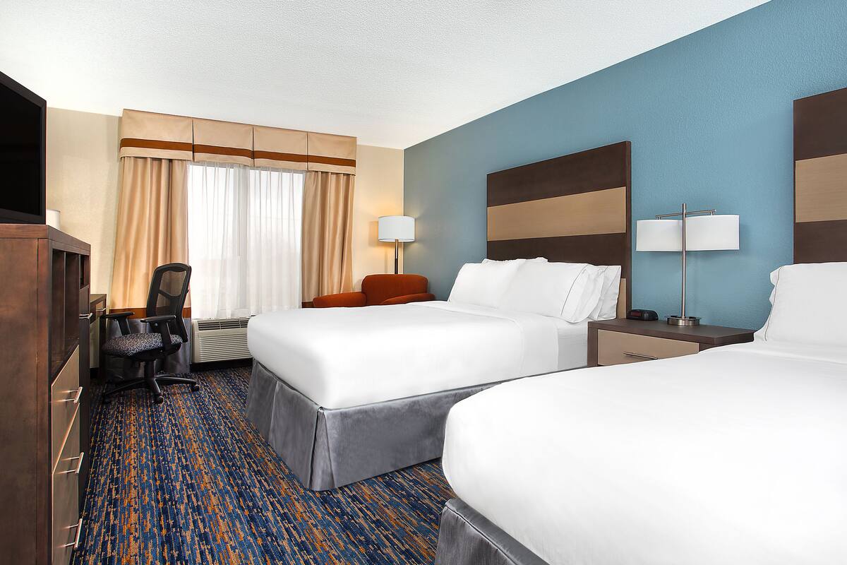 A hotel room with two beds, a desk, a chair, a lamp, and a large window with curtains.