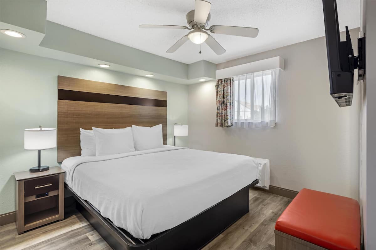 A hotel room with a bed, nightstand, lamp, ceiling fan, window with curtains, and a red bench.