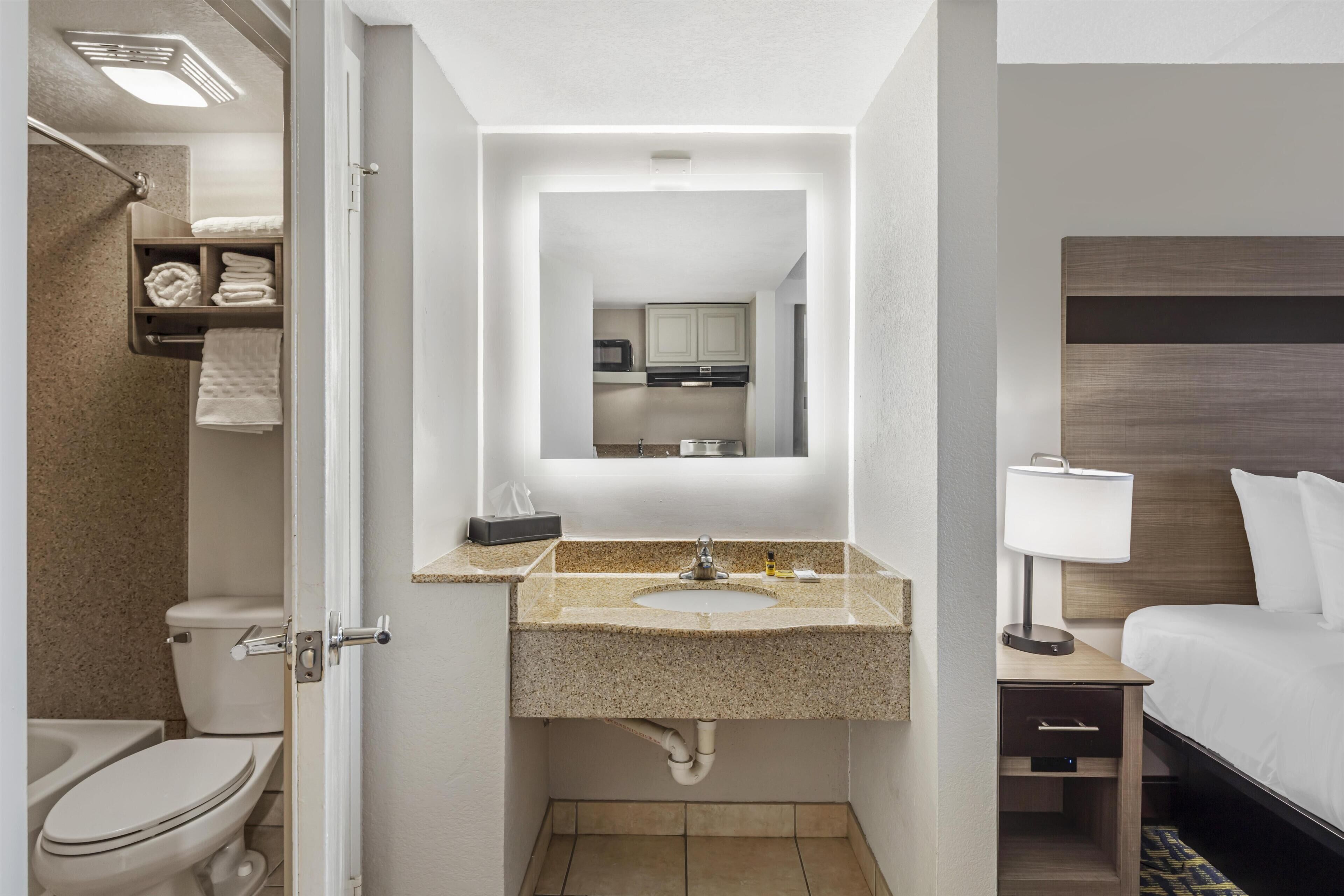 Un baño de hotel con inodoro, lavamanos, espejo y una cama con mesita de noche.