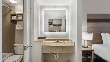 Un baño de hotel con inodoro, lavamanos, espejo y una cama con mesita de noche.
