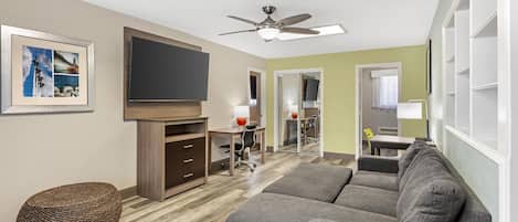 A living room with a grey sofa, a flat-screen TV, a ceiling fan, and a woven ottoman.
