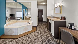 A hotel room with a large mirror, a bathtub, a desk with a chair, and a kitchenette.