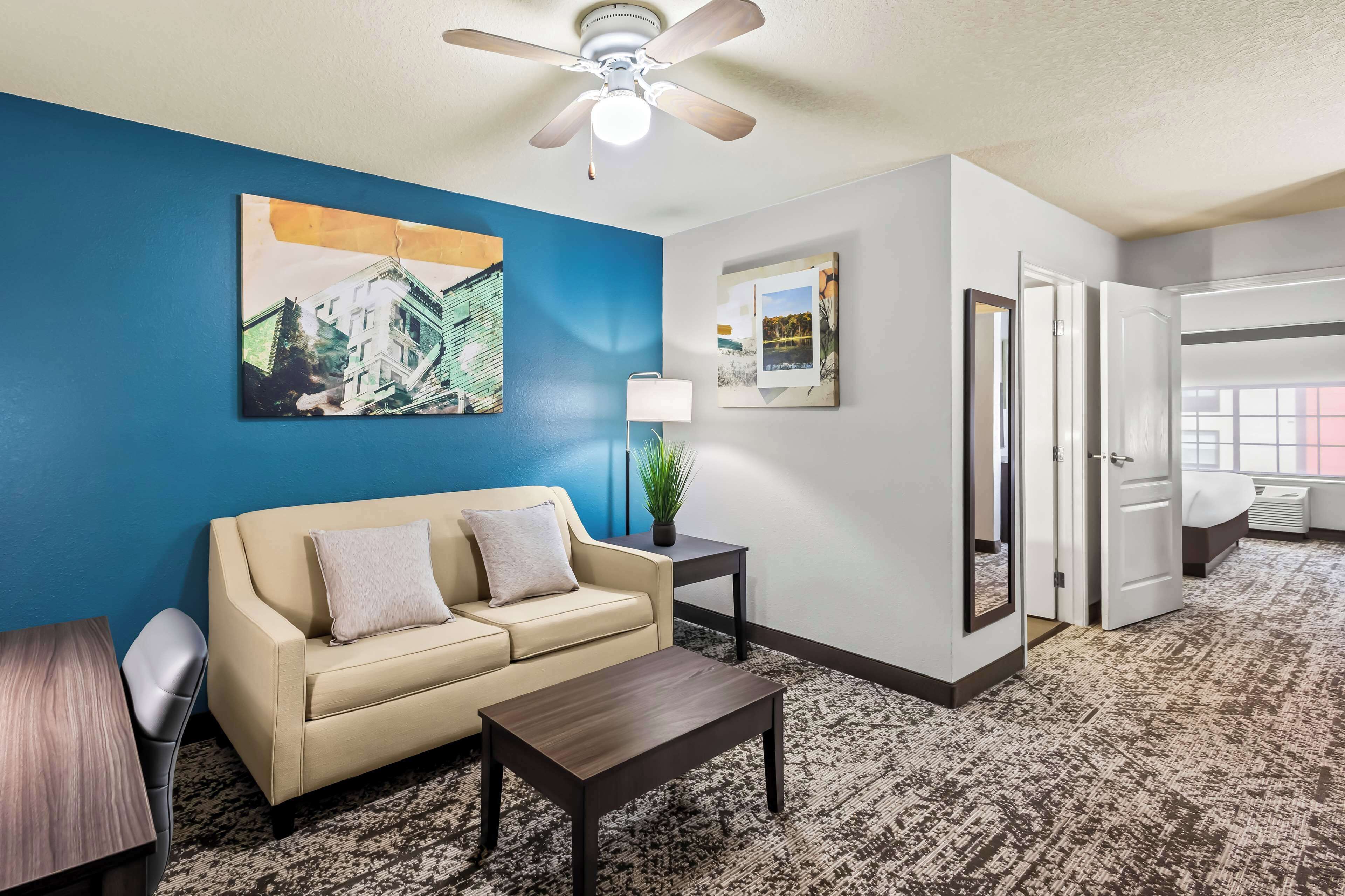 A hotel room with a blue accent wall, a beige sofa, a small table, a ceiling fan, and a bed with a white headboard.