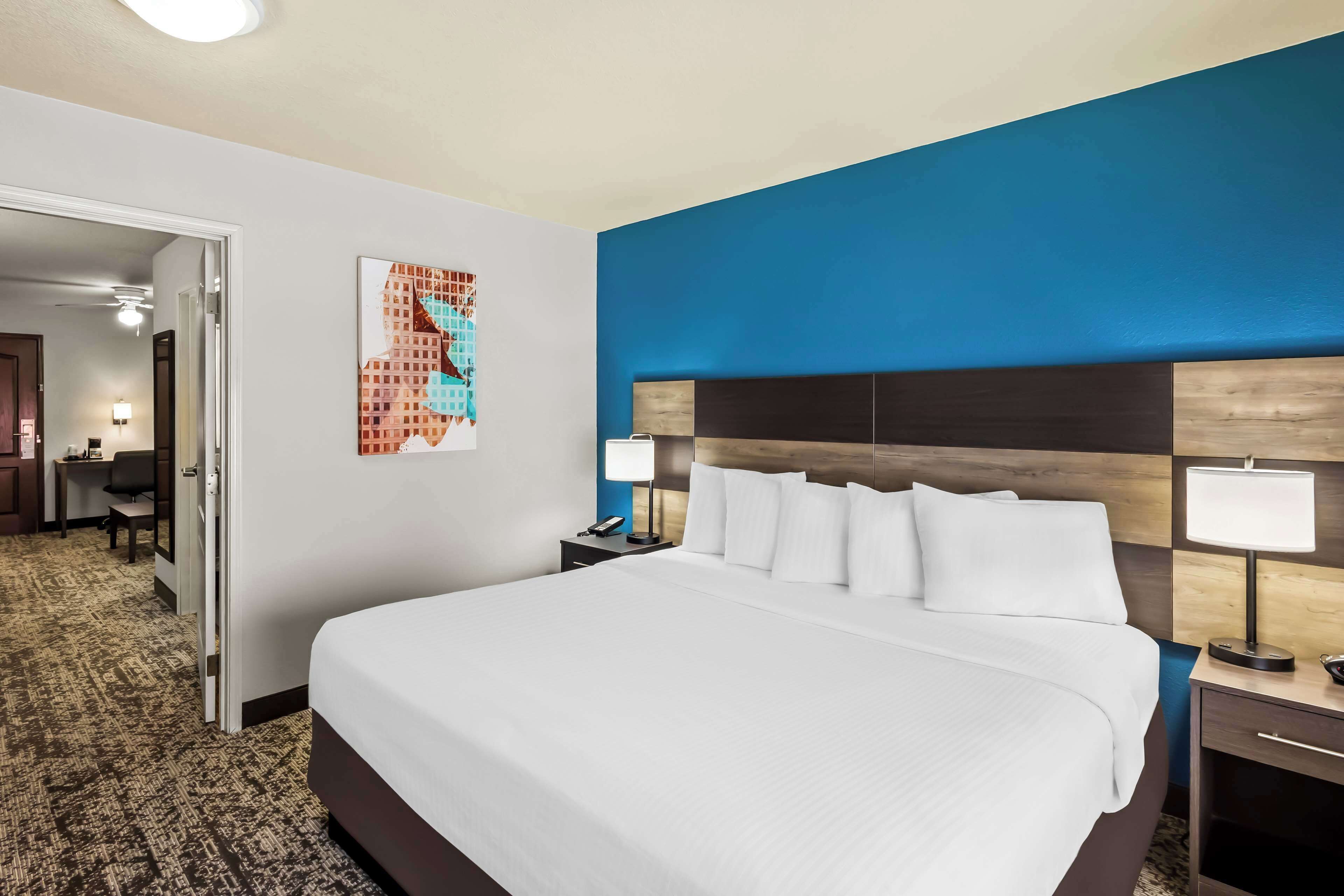 A hotel room with a large bed, a desk, a chair, a nightstand with a lamp, and a wall art piece.