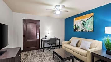 A hotel room with a dark wood door, a beige sofa, a desk with a chair, a flat-screen TV, a ceiling fan, and a painting on the wall.
