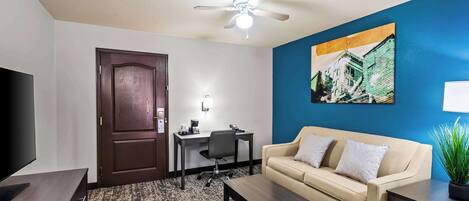 A hotel room with a dark wood door, a beige sofa, a desk with a chair, a flat-screen TV, a ceiling fan, and a painting on the wall.