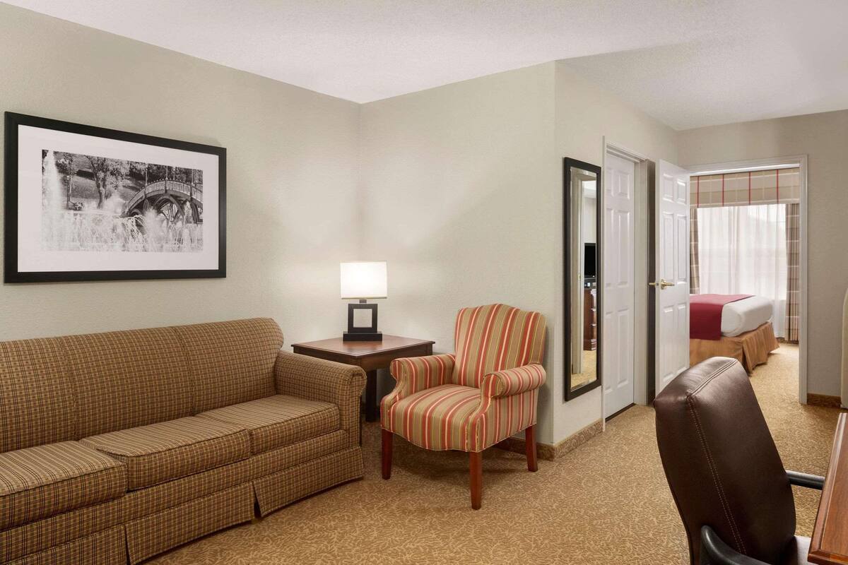 A hotel room with a plaid sofa, a striped armchair, a small table with a lamp, a framed picture on the wall, and a door leading to a bedroom with a bed and a window.