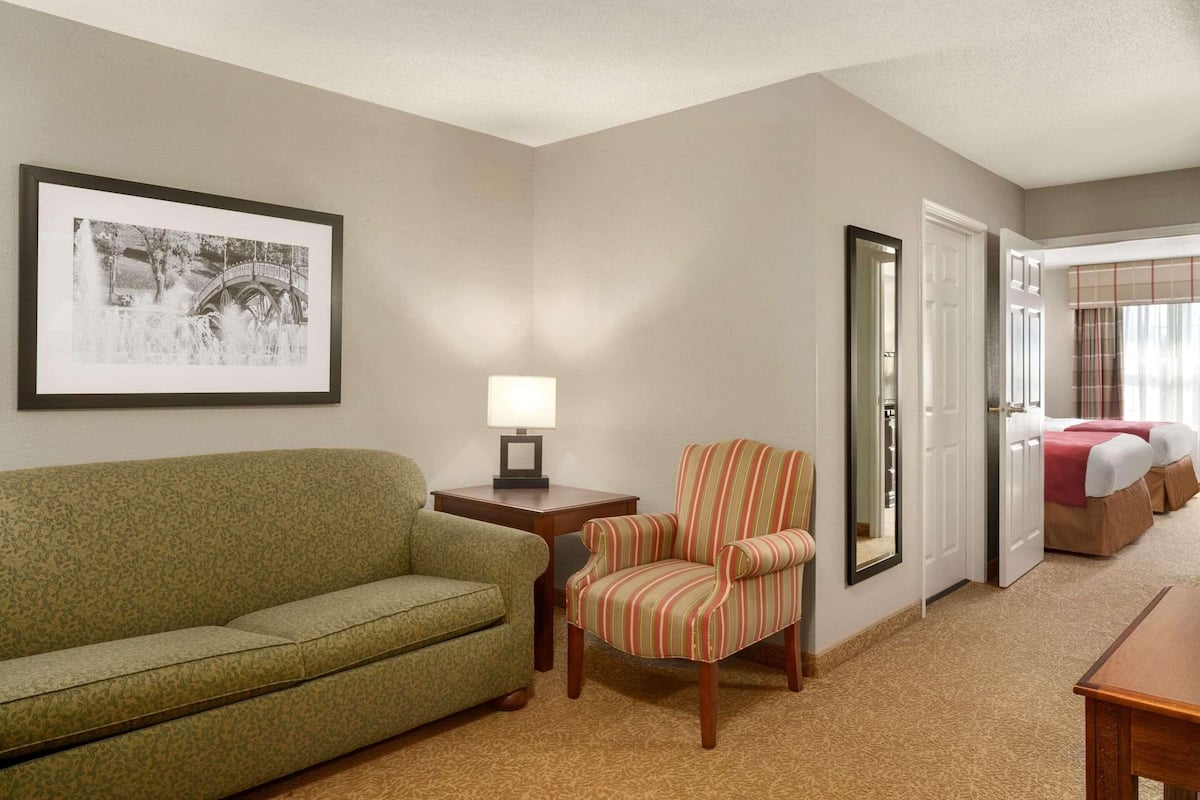 A hotel room with a green sofa, a striped armchair, a framed picture on the wall, a bedside table with a lamp, and a bed with a red blanket.