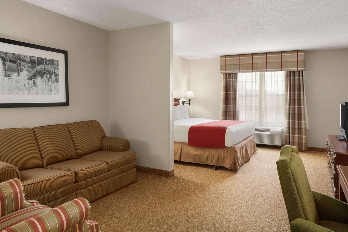 A hotel room with a bed, a sofa, a chair, a lamp, a television, and a framed picture on the wall.