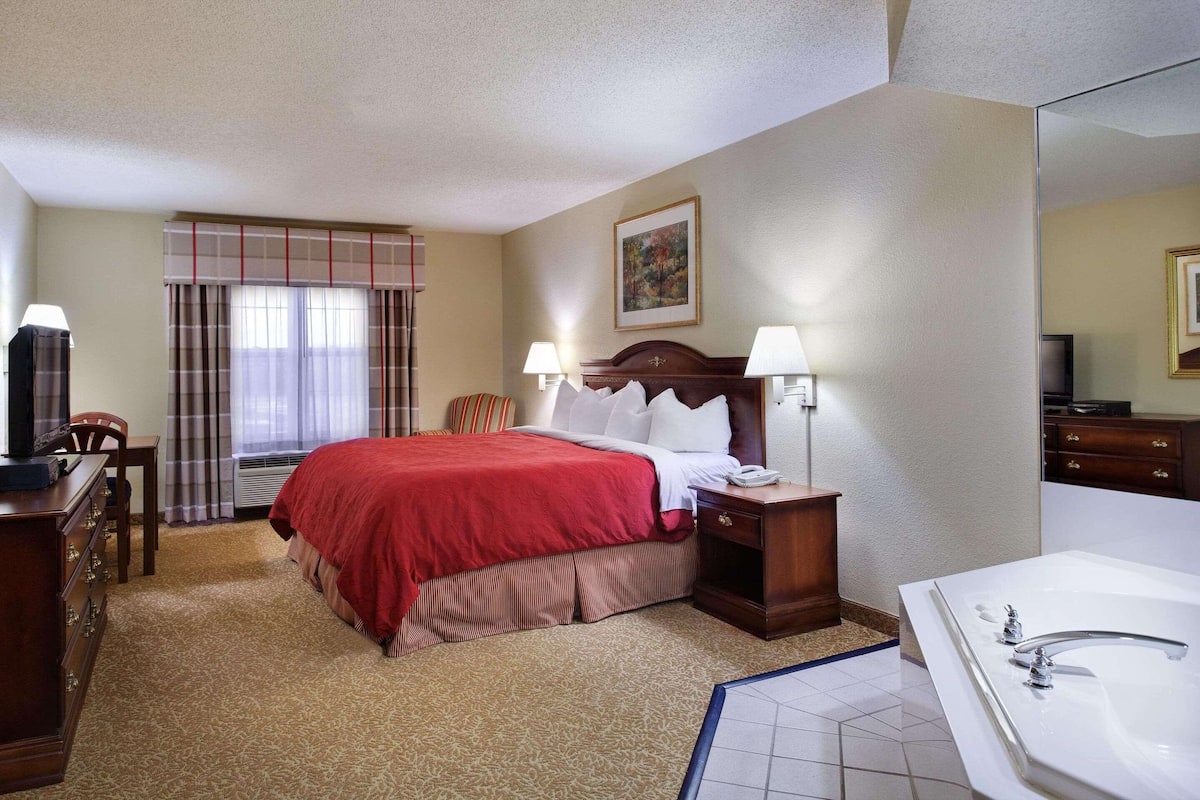 A hotel room with a bed, bedside table, lamp, TV, and a large bathroom with a bathtub.