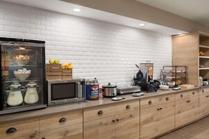 Una cocina moderna con horno empotrado, microondas y diversos electrodomésticos. Hay armarios de madera y una encimera con alimentos y una cafetera.
