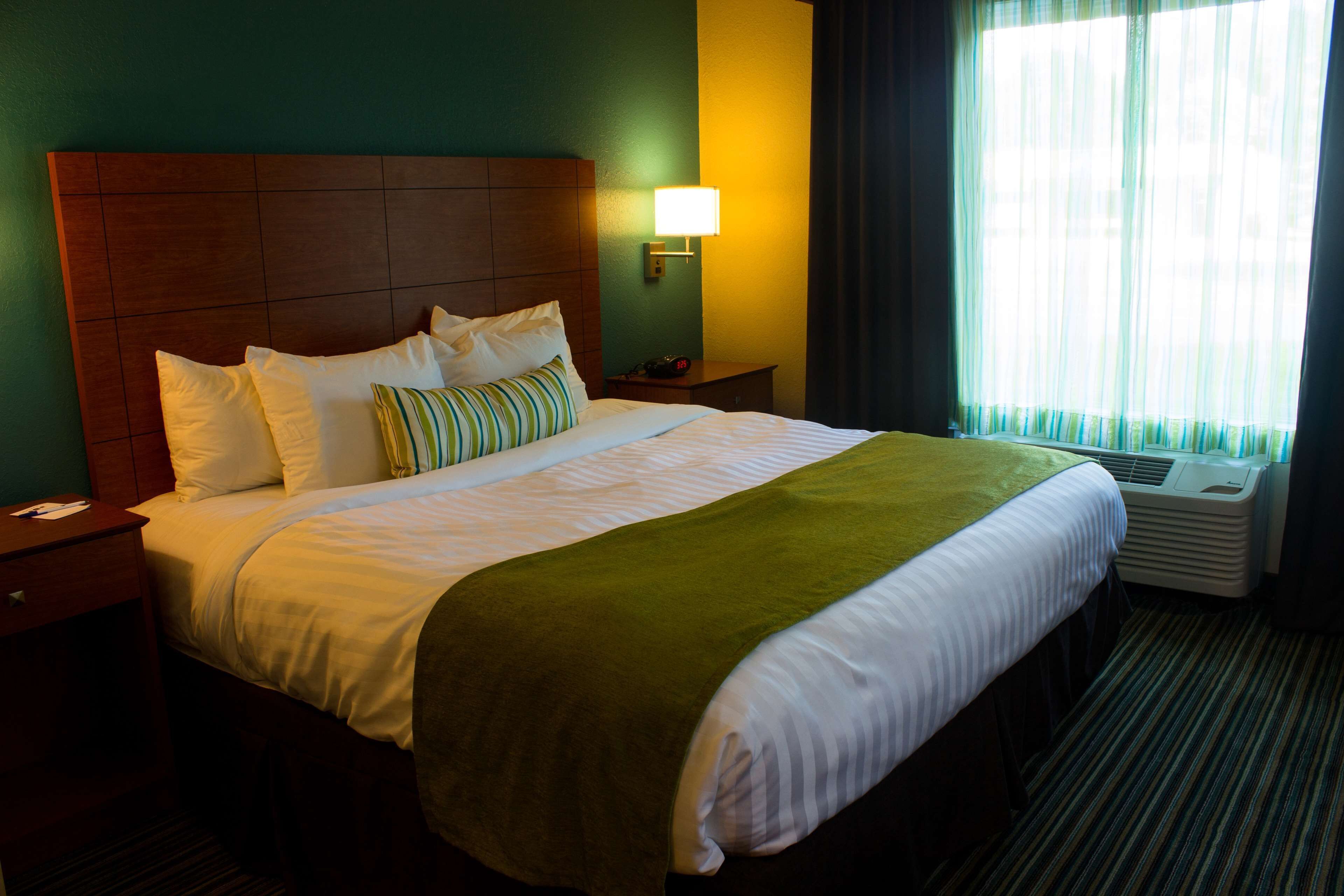 Habitación de hotel con una cama grande, una mesita de noche, una lámpara y un ventanal con cortinas.