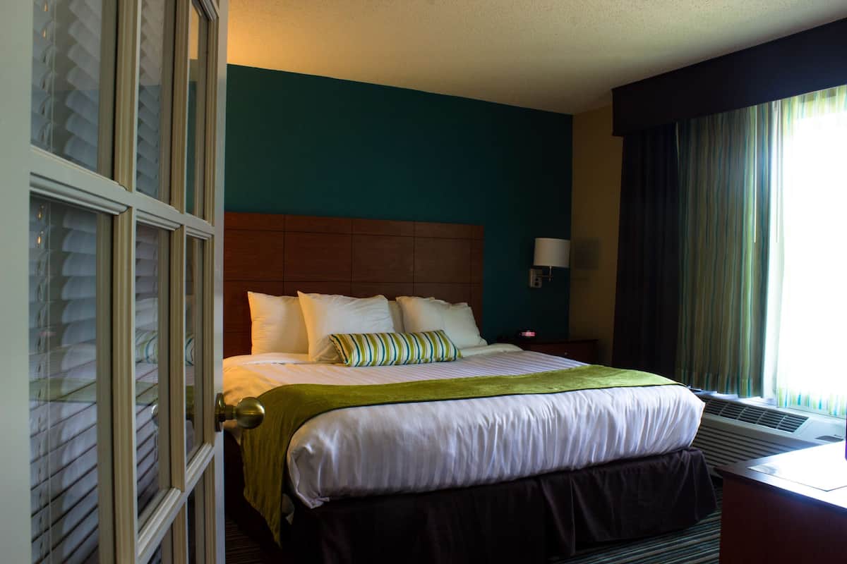 Una habitación de hotel con cama, mesita de noche, lámpara y ventana con cortinas.