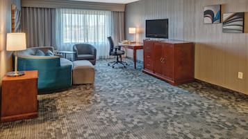 A hotel room with a sofa, armchair, desk, television, and artwork on the wall.