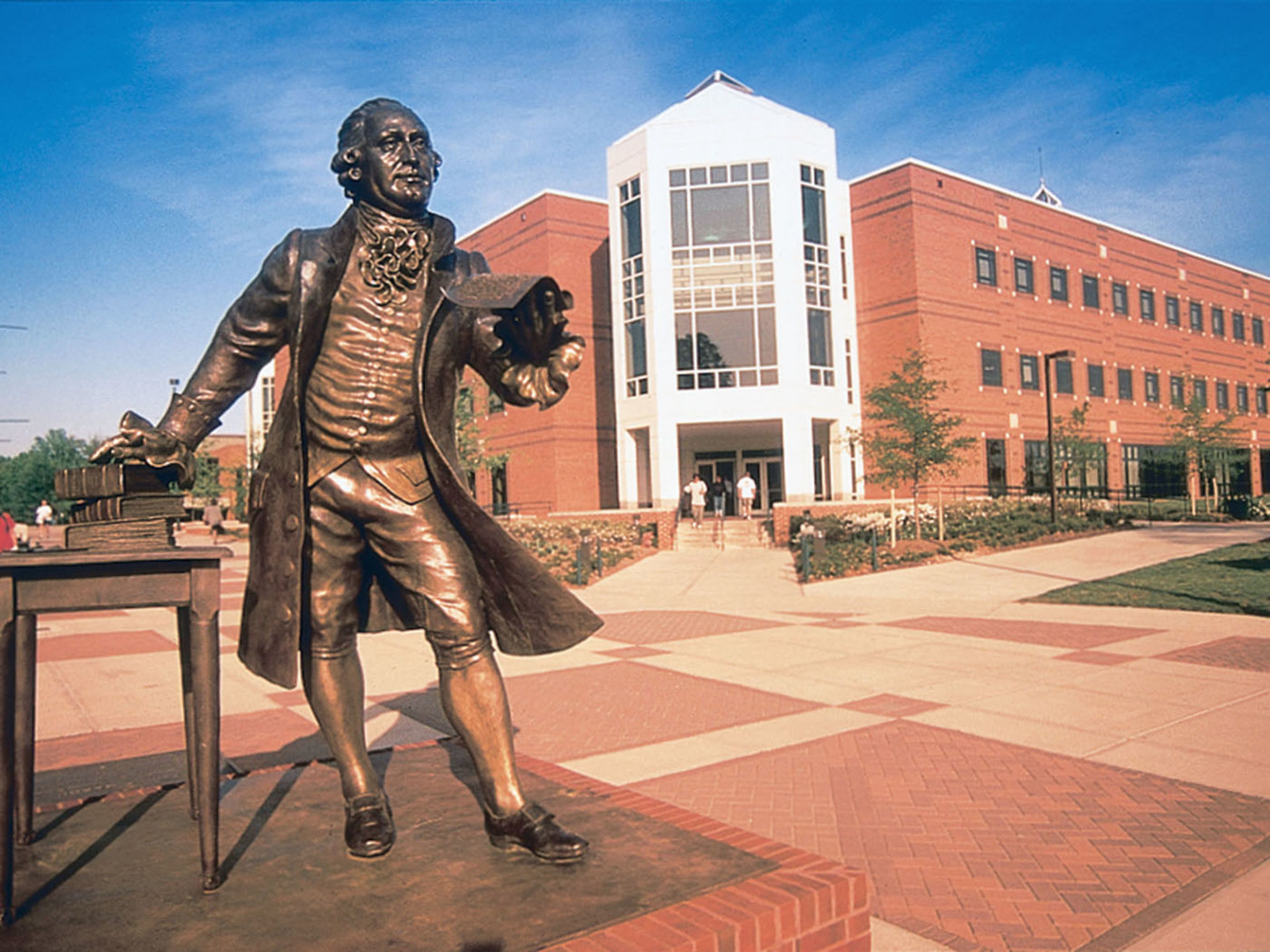 A bronze statue of a historical figure in a formal coat and hat, positioned on a brick-paved area with a modern building in the background.