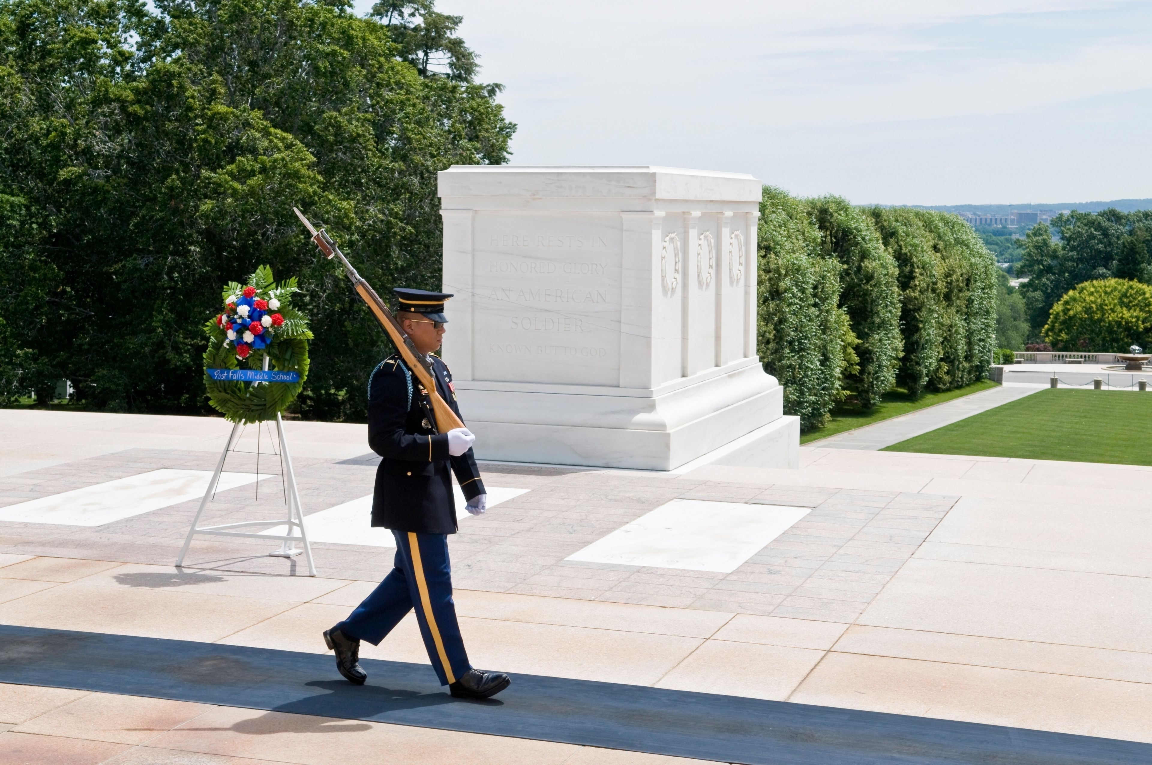 A uniformed guard carrying a rifle, a wreath, and a flagpole with a wreath in a formal setting with a monument and trees in the background.