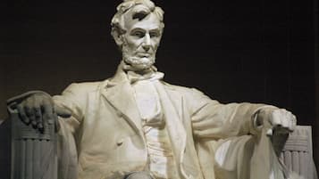 Une statue représentant une figure assise, vêtue d’un drapé, assise sur un fauteuil à accoudoirs, le tout sur un fond sombre.