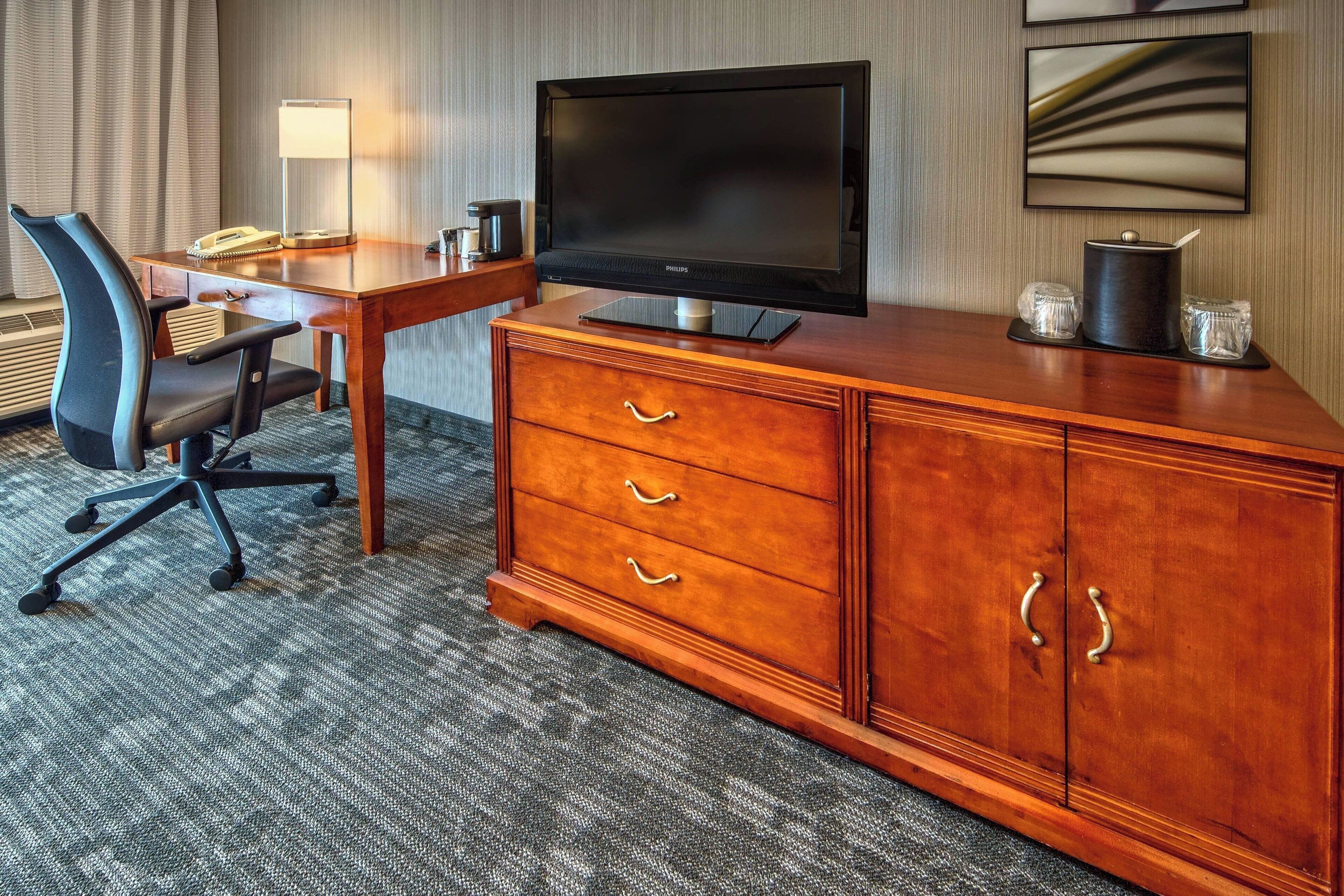 A hotel room with a wooden desk, a black flat-screen TV, a chair, a lamp, and a coffee maker.