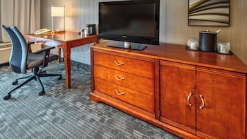 A hotel room with a wooden desk, a black flat-screen TV, a chair, a lamp, and a coffee maker.