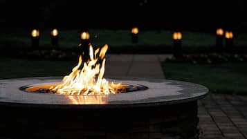 Un foyer circulaire en pierre, avec des flammes, entouré d’un muret en pierre et d’un chemin pavé en briques.