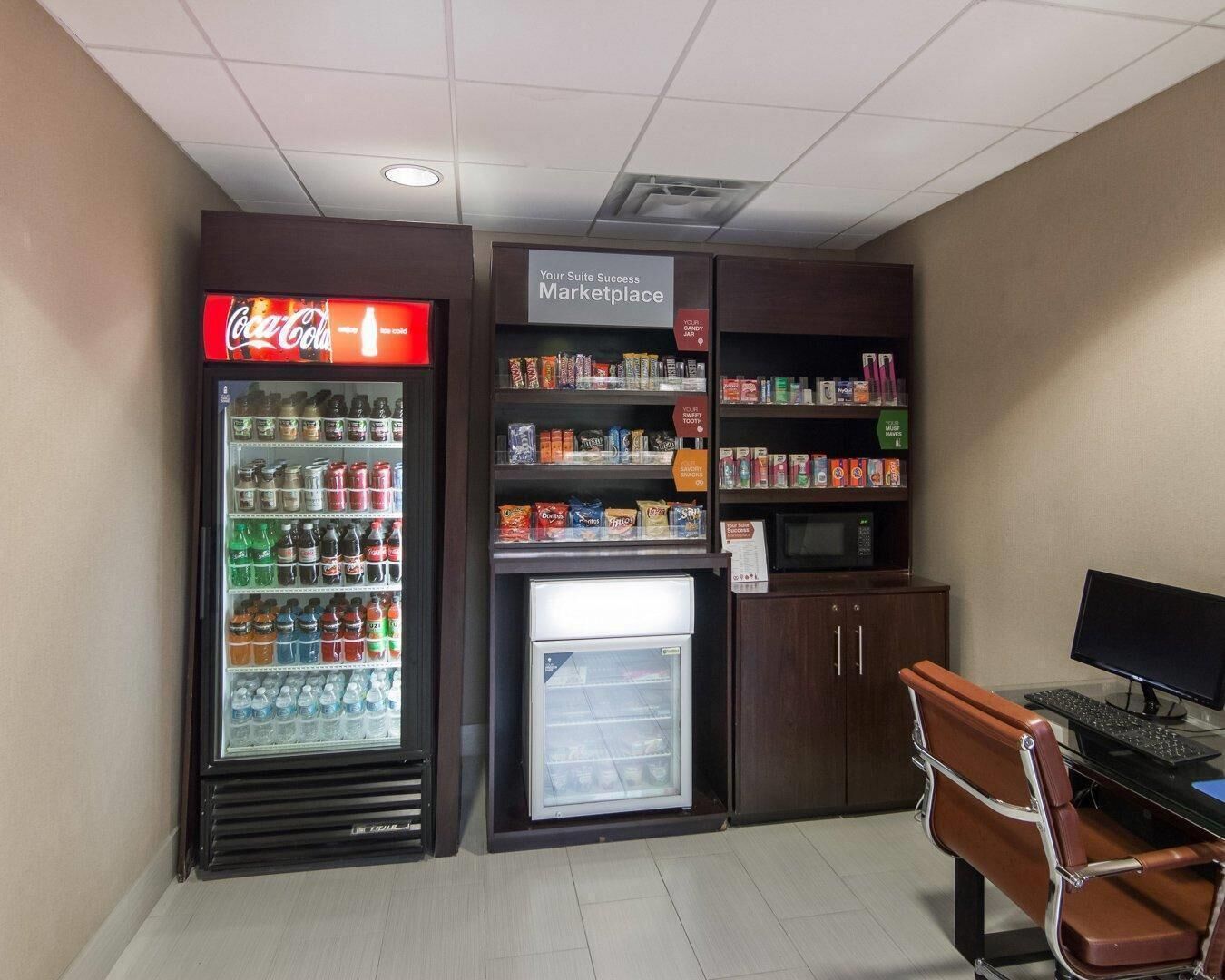 A vending machine area with a Coca-Cola cooler and a refrigerated section, a small desk with a computer, and a chair.