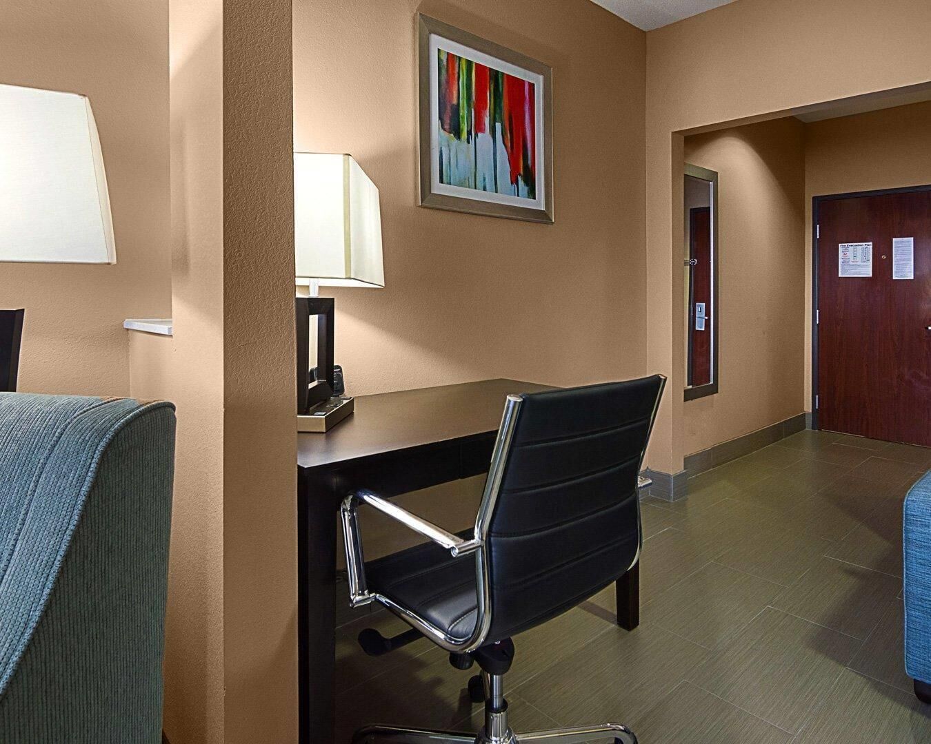 Una habitación de hotel con escritorio, silla, lámpara, obras de arte y una puerta.