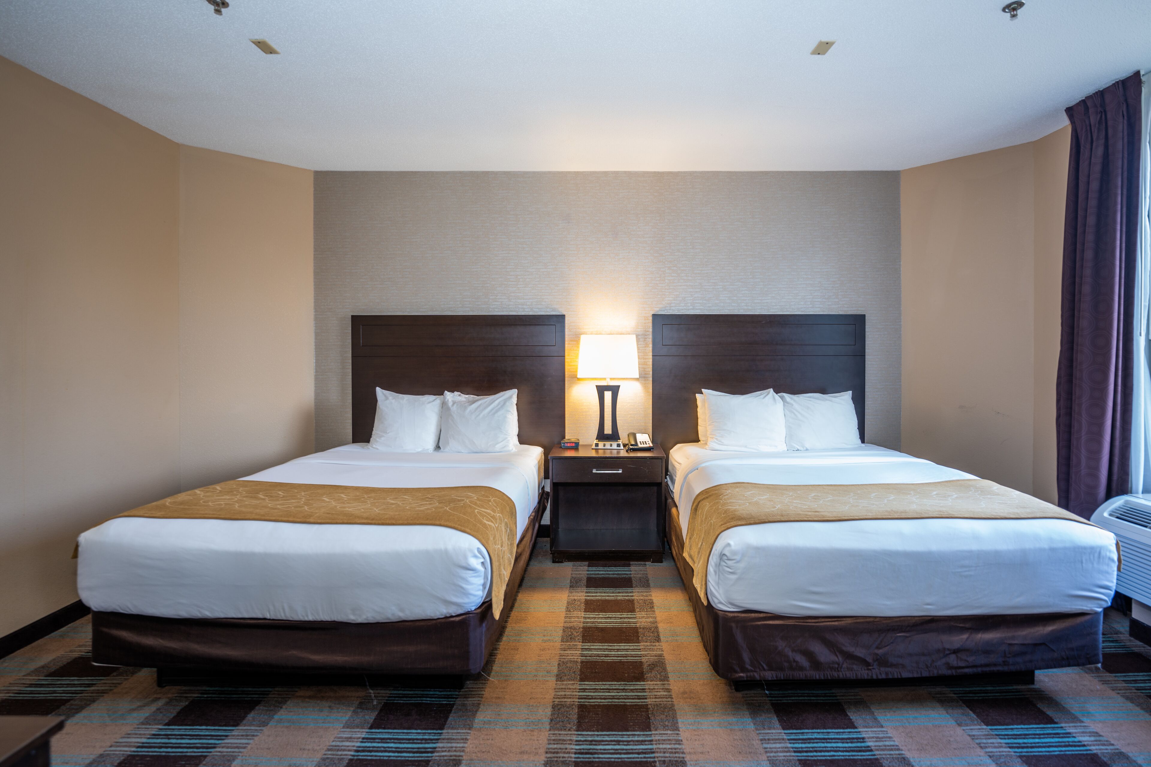 A hotel room with two beds, a nightstand, a lamp, and a window with curtains.
