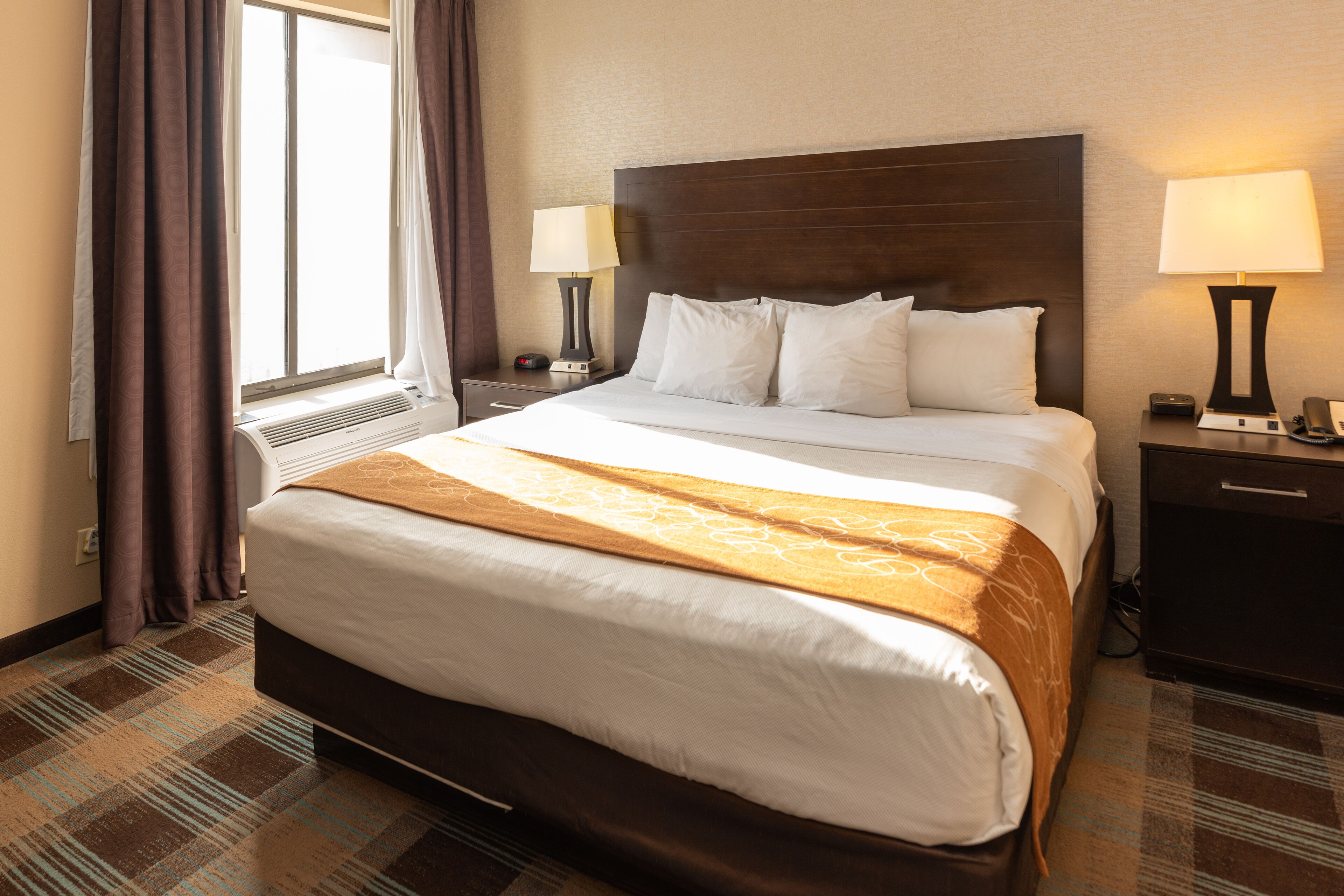 Hotellihuone, jossa on suuri sänky, kaksi yöpöytää valaisimineen, ikkuna verhoilla ja matolla peitetty lattia.