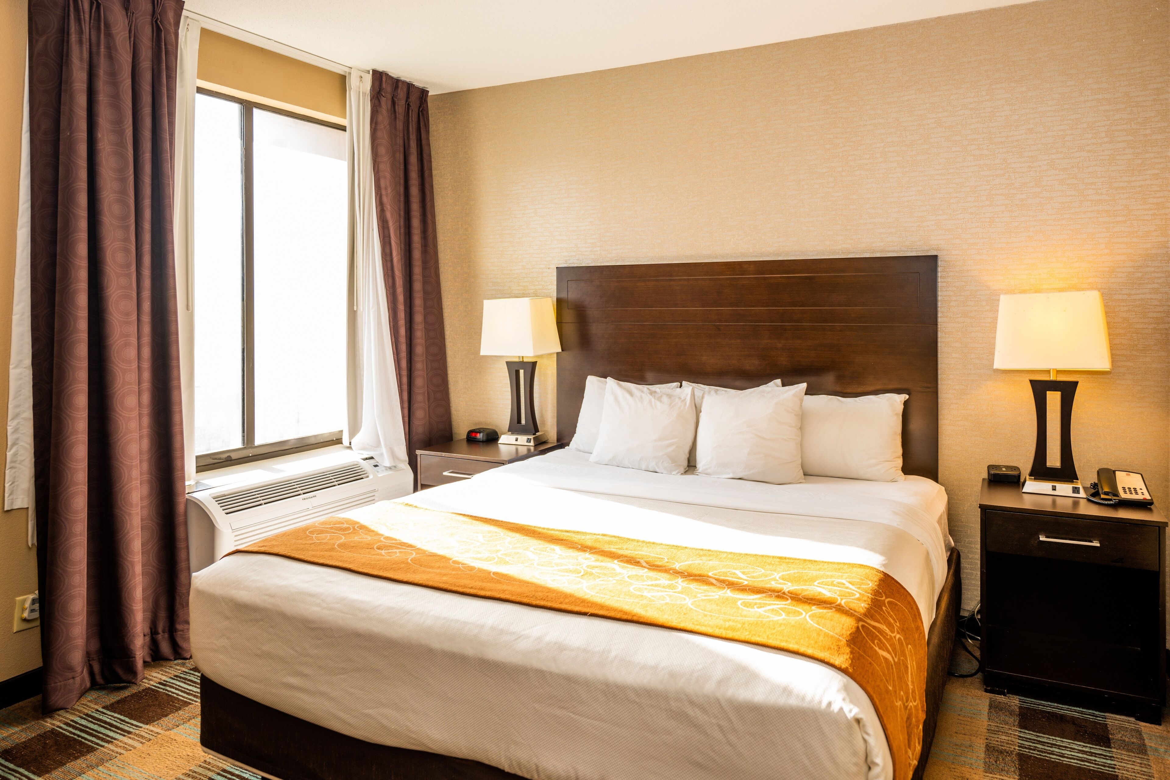 A hotel room with a large bed, two bedside tables with lamps, a window with curtains, and a wall-mounted air conditioning unit.