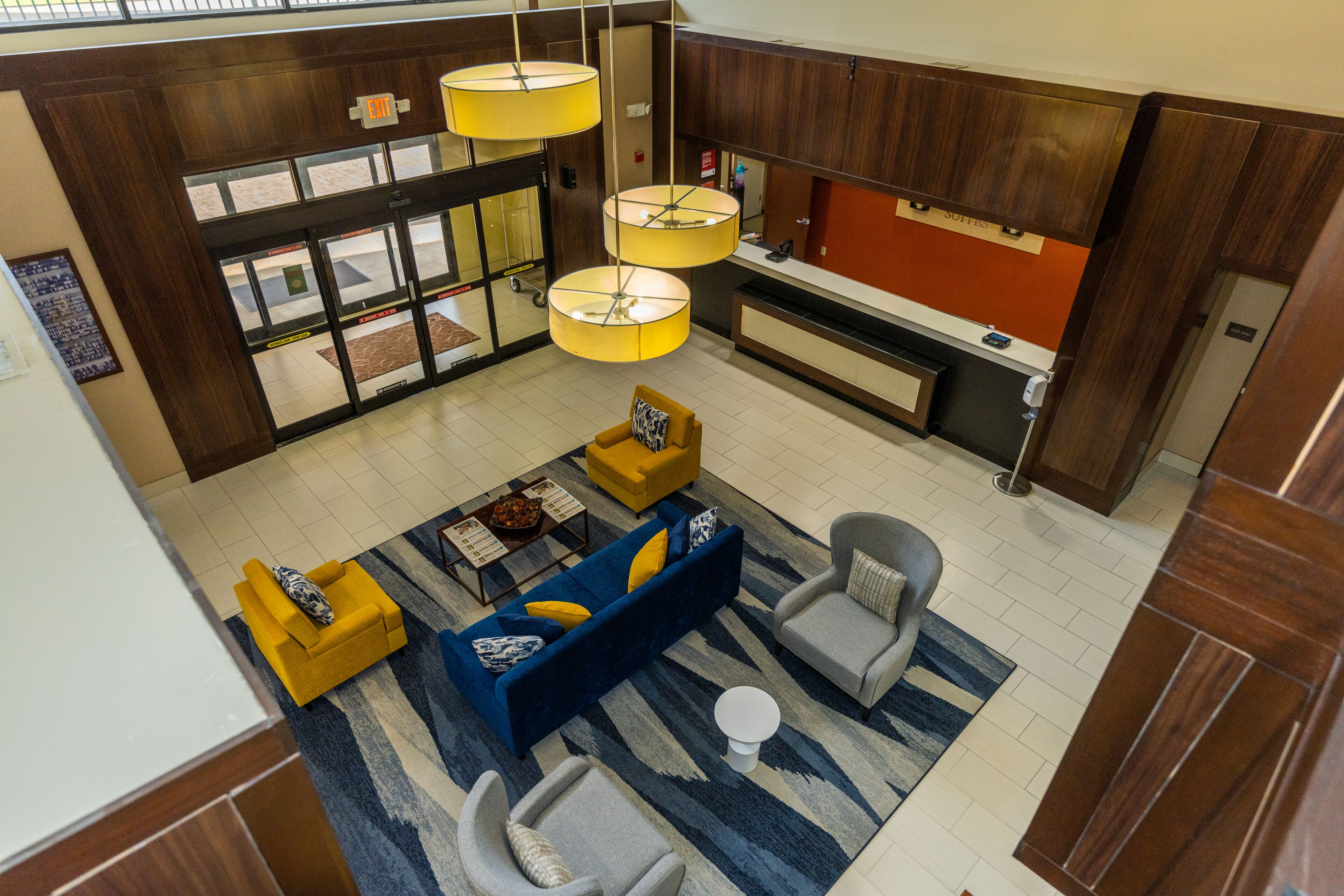 Un vestíbulo amplio con un sofá azul, sillones amarillos y un sillón gris. Se observan tres lámparas colgantes, un mostrador de recepción y un ventanal con vistas.