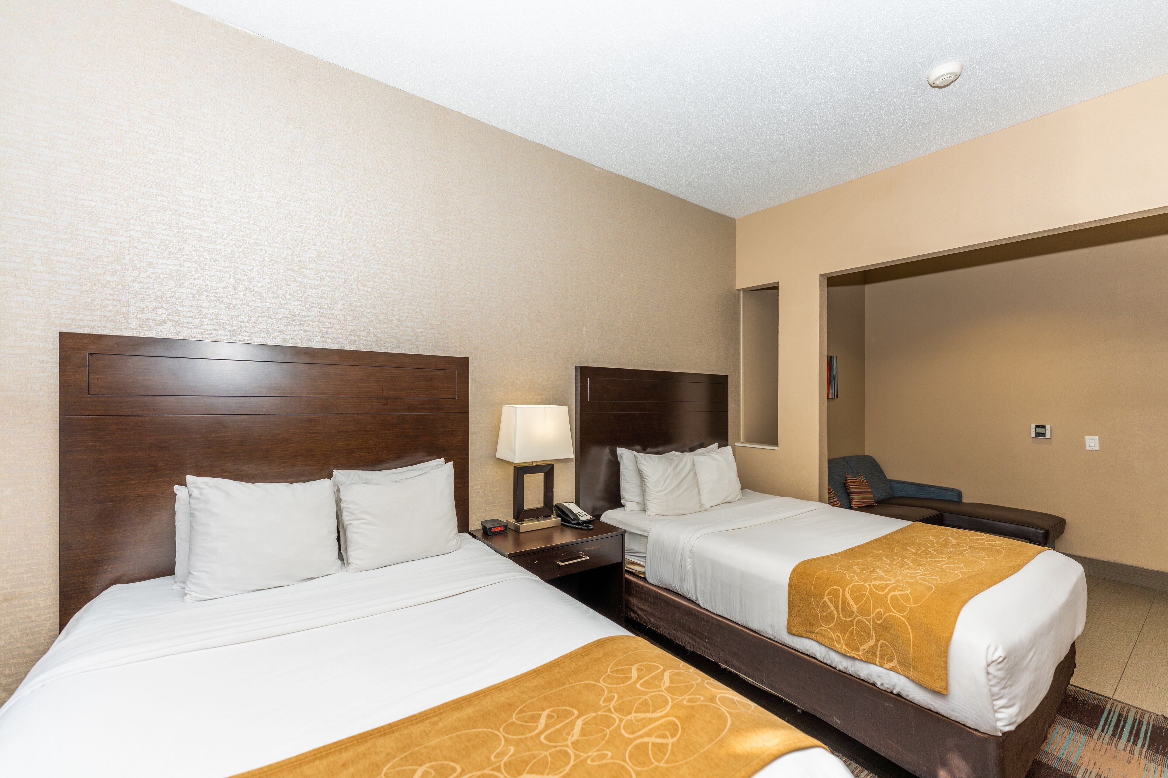 A hotel room with two beds, a nightstand with a lamp, and a seating area with a chair.