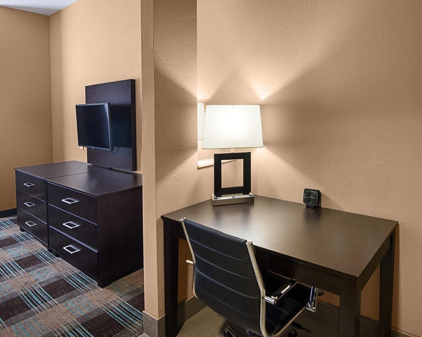 A hotel room with a desk, chair, television, and a lamp.