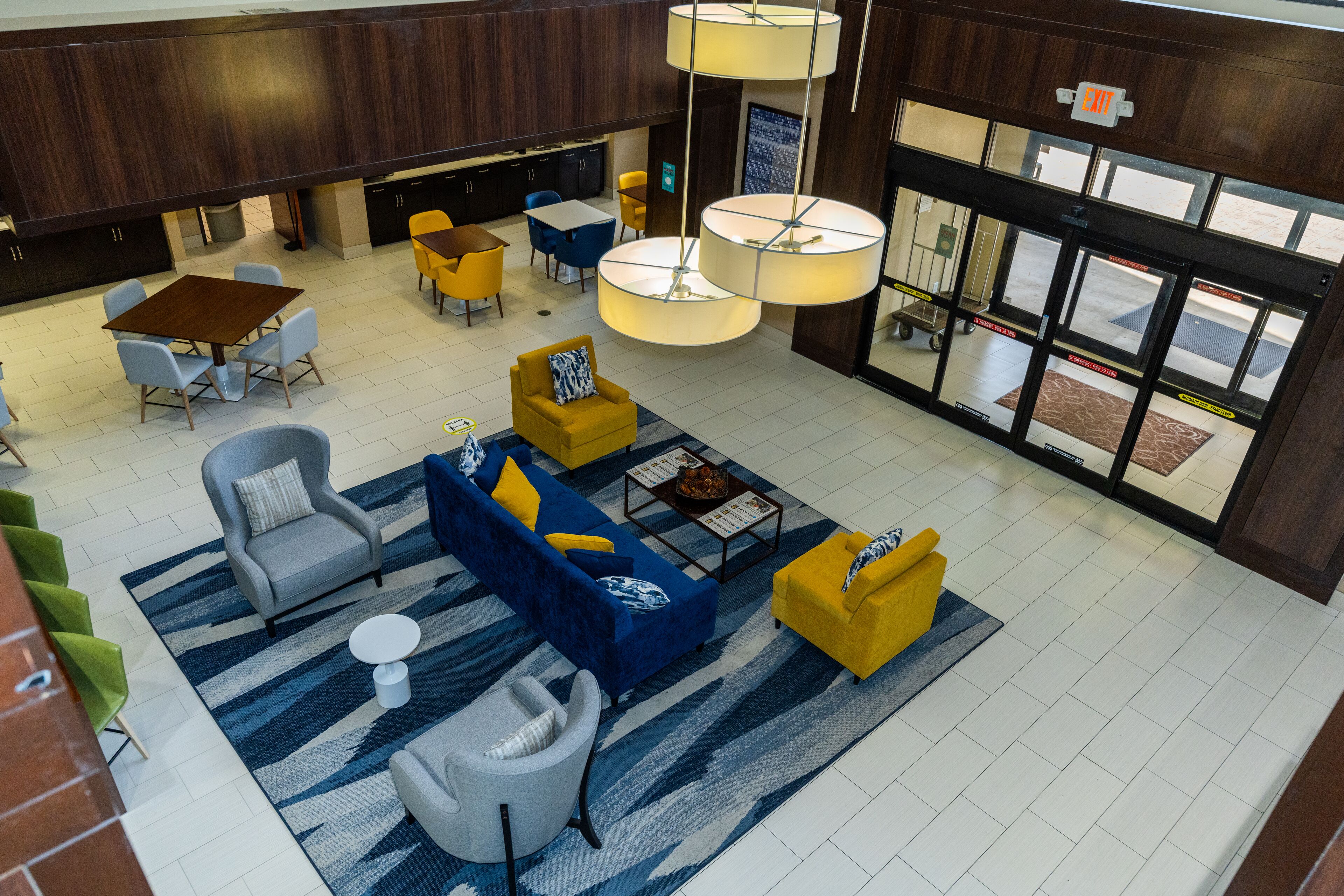 A spacious lobby with a central seating area featuring sofas and armchairs, a coffee table, and a rug. There are multiple tables and chairs around the seating area, and a large window providing natural light.