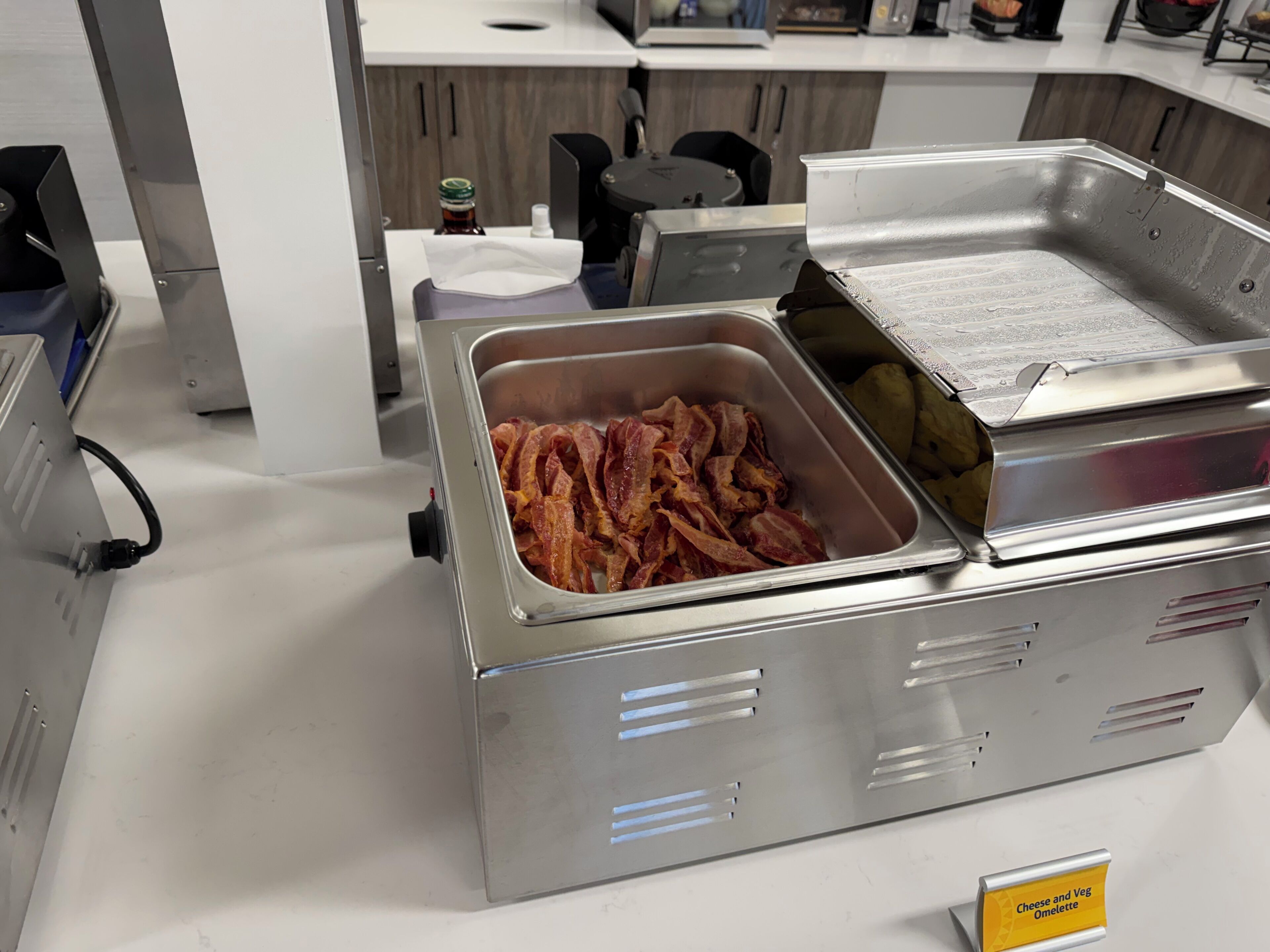 A buffet station with a container of bacon and a sign indicating 'Cheese and Veg Omelette.'