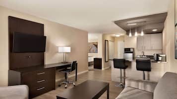 A modern hotel room with a flat-screen TV, a desk, a chair, a sofa, and a kitchenette in the background.