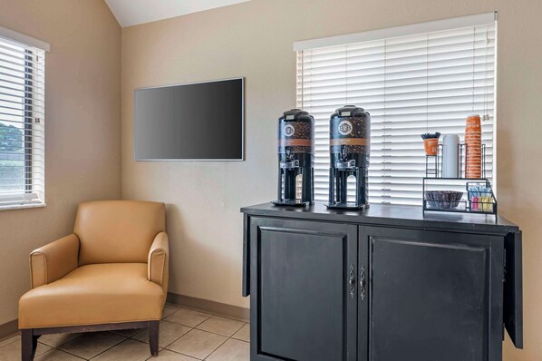 A room with a beige armchair, a black cabinet with coffee makers, and a large window with blinds.