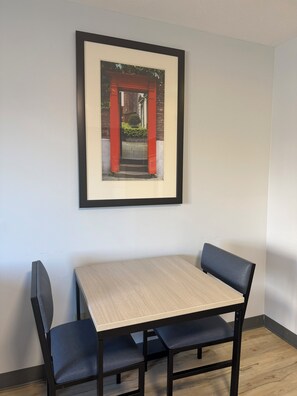 Un cuadro enmarco de un arcojón rojo en una pared, una mesa pequeña con dos sillas y un suelo de madera.