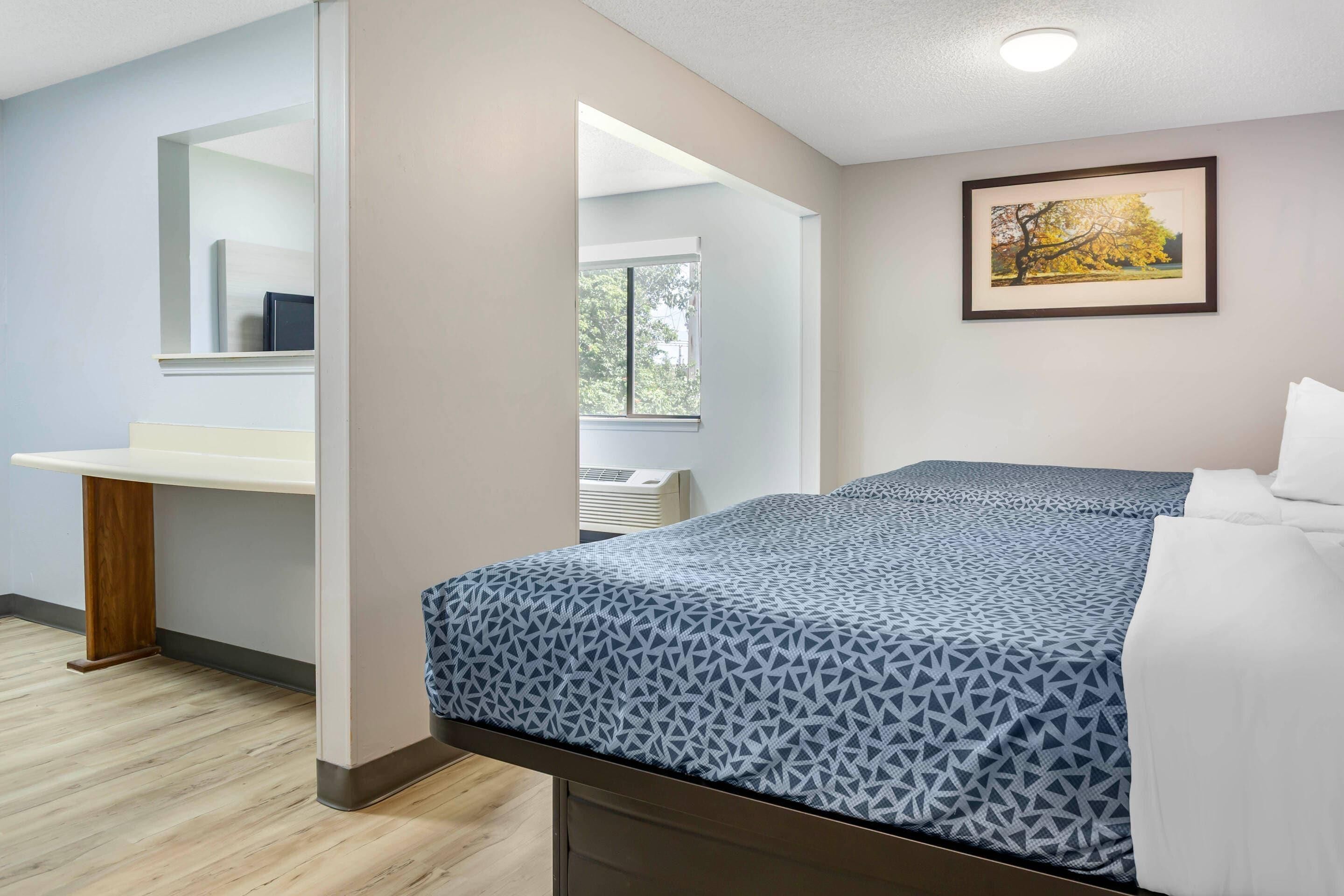 A hotel room with a bed, a bedside table, a mirror, a window, and a framed picture on the wall.