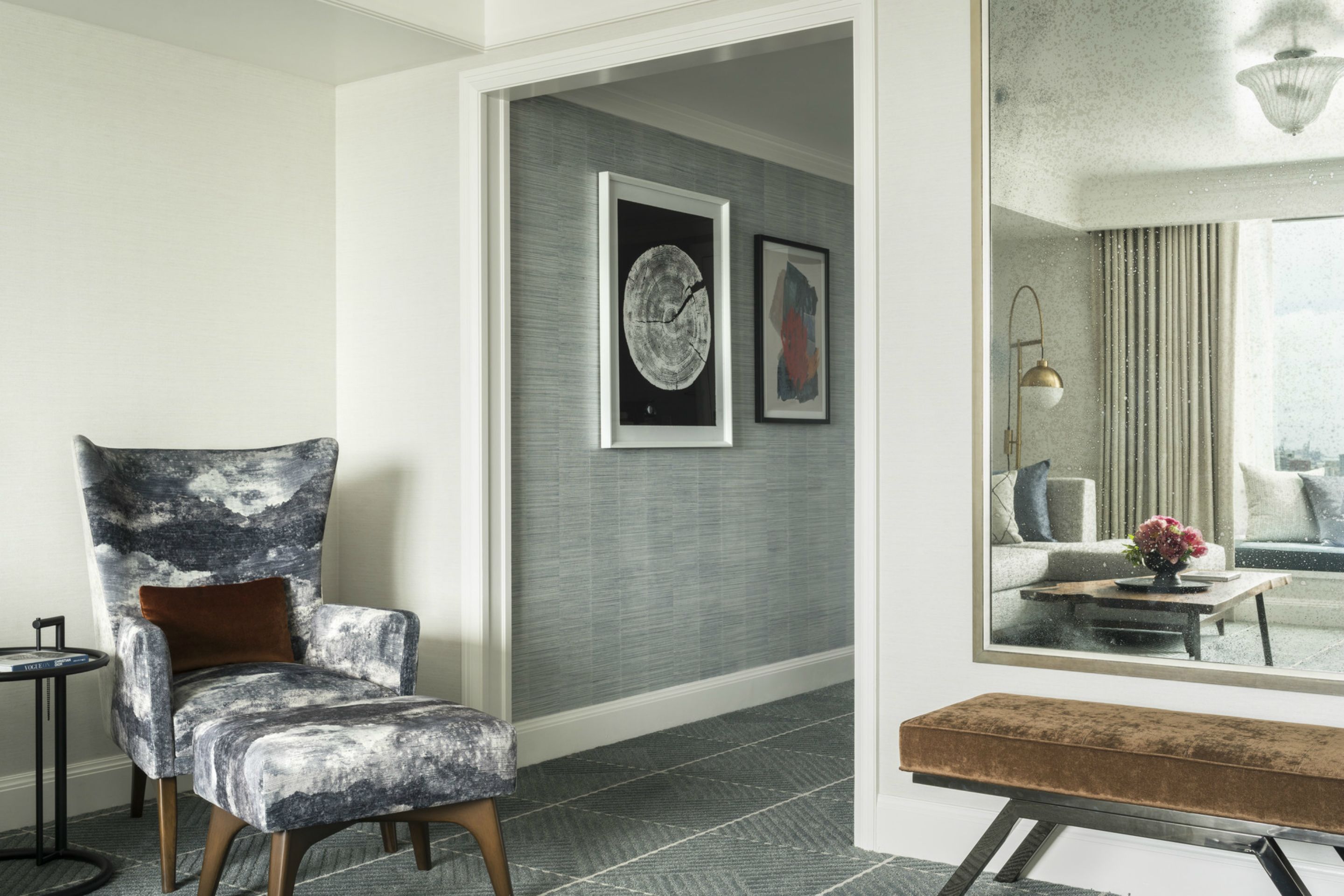 Een moderne woonkamer met een grote fauteuil, een bankje en een spiegel die een bank en een raam weerspiegelt.
