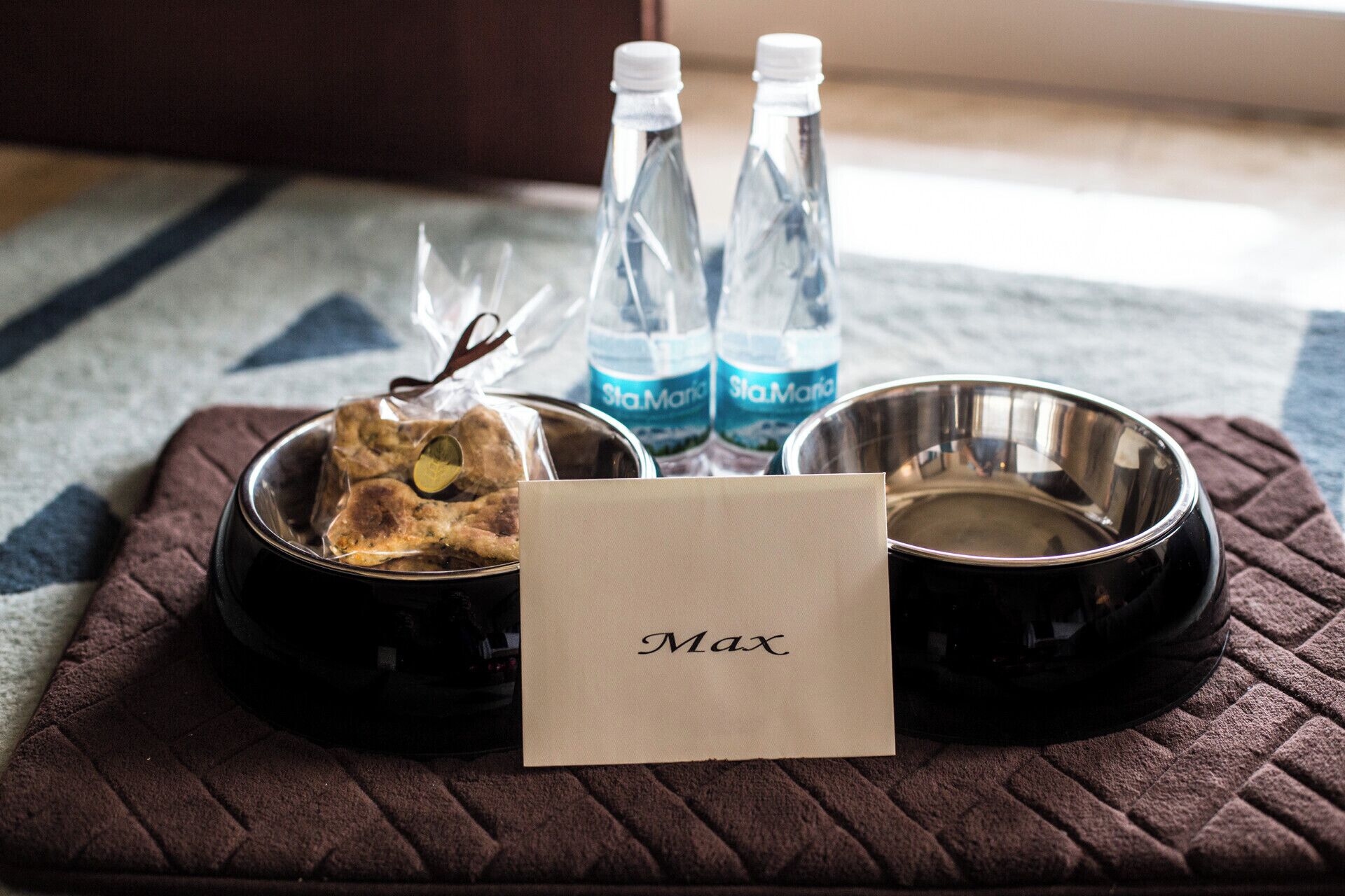 A pet bed with a name tag, two water bottles, and a bowl.