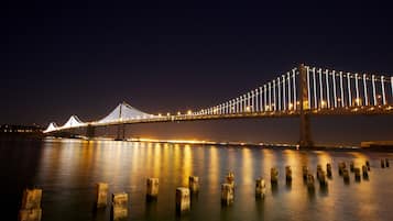 A nighttime view of a large bridge with illuminated cables and a lit roadway, spanning over a calm body of water with several concrete pillars in the foreground.