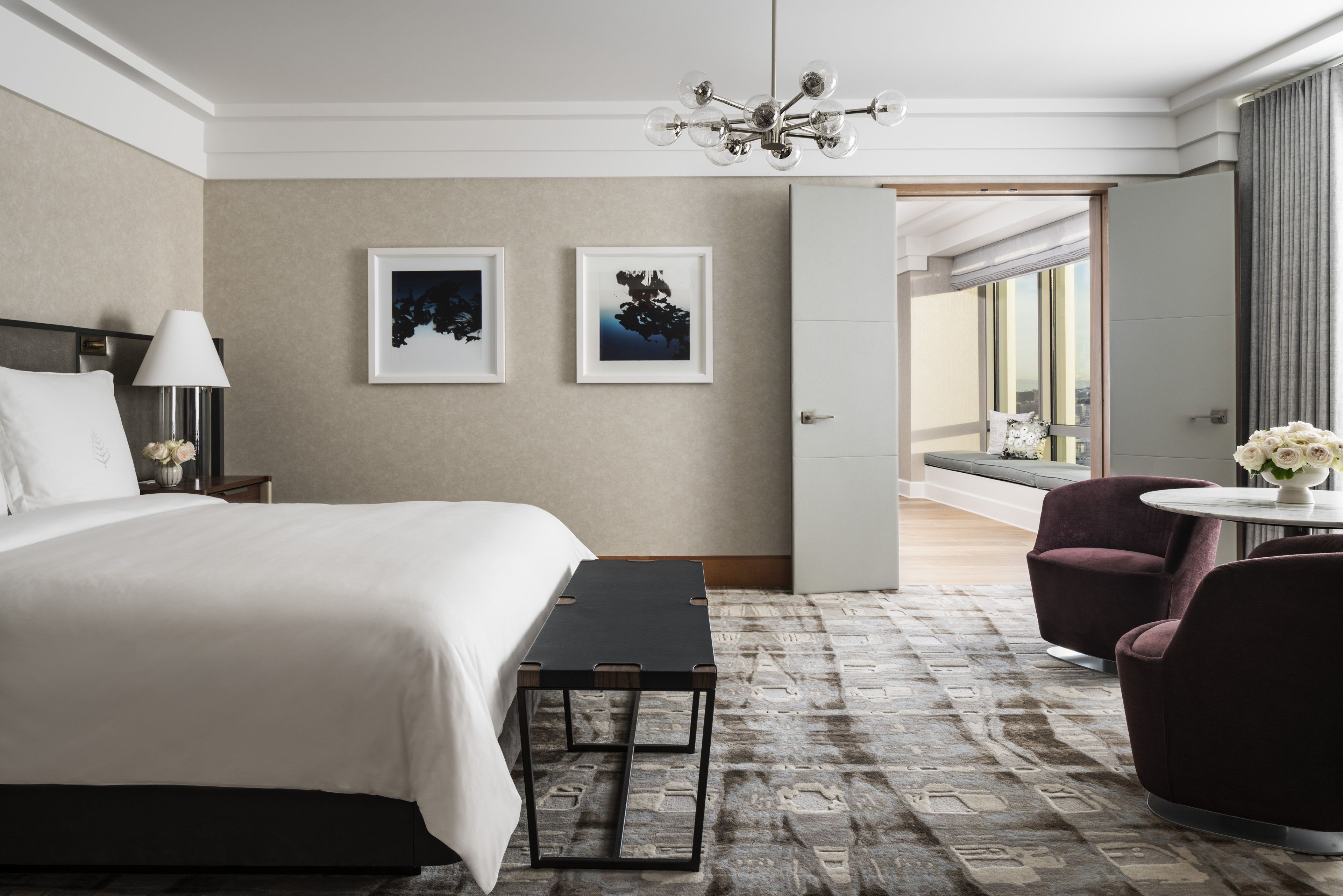 Habitación de hotel con una cama grande, una mesita pequeña, dos sillas, un candelabro y cuadros en la pared.